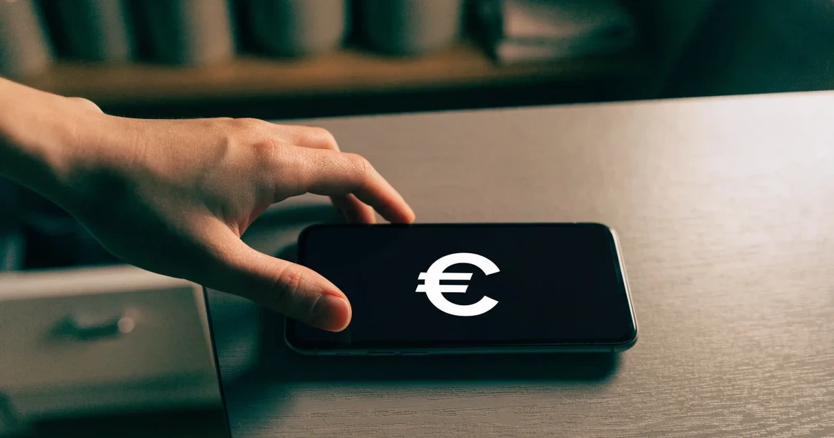 Eine Hand legt ein paar Münzen neben ein umgedrehtes Smartphone auf eine graue Tischplatte, dahinter verschwommen ein Küchenregal.