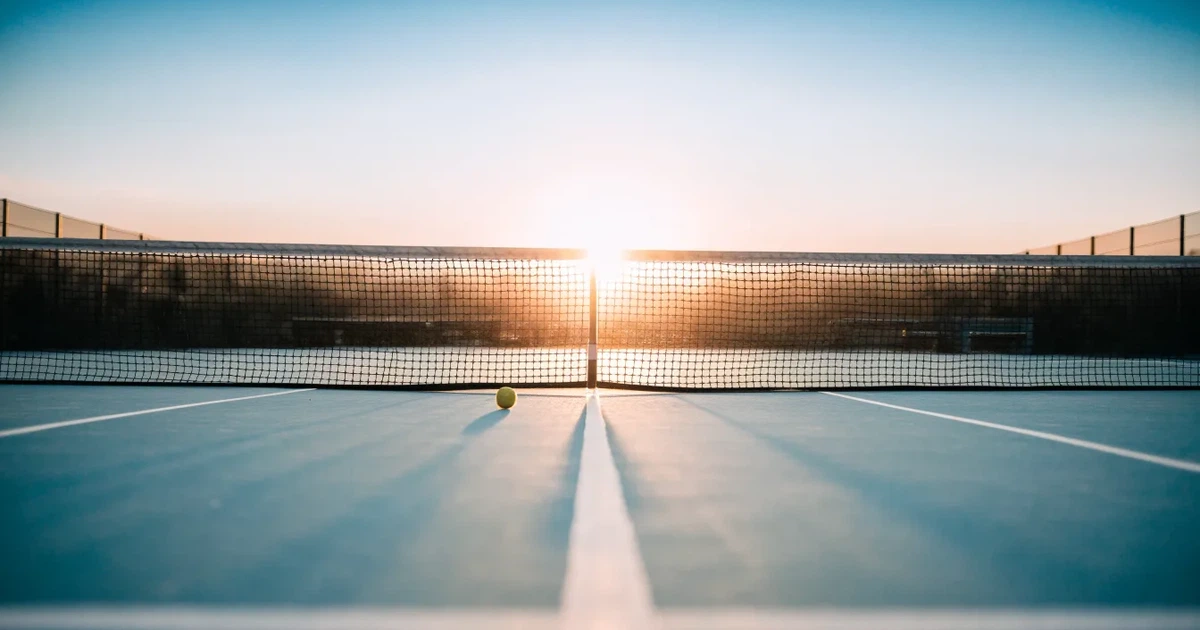 Ein leerer Tennisplatz mit Hartplatzbelag in der Morgendämmerung, das Netz im Gegenlicht und lange Schatten der Linien.