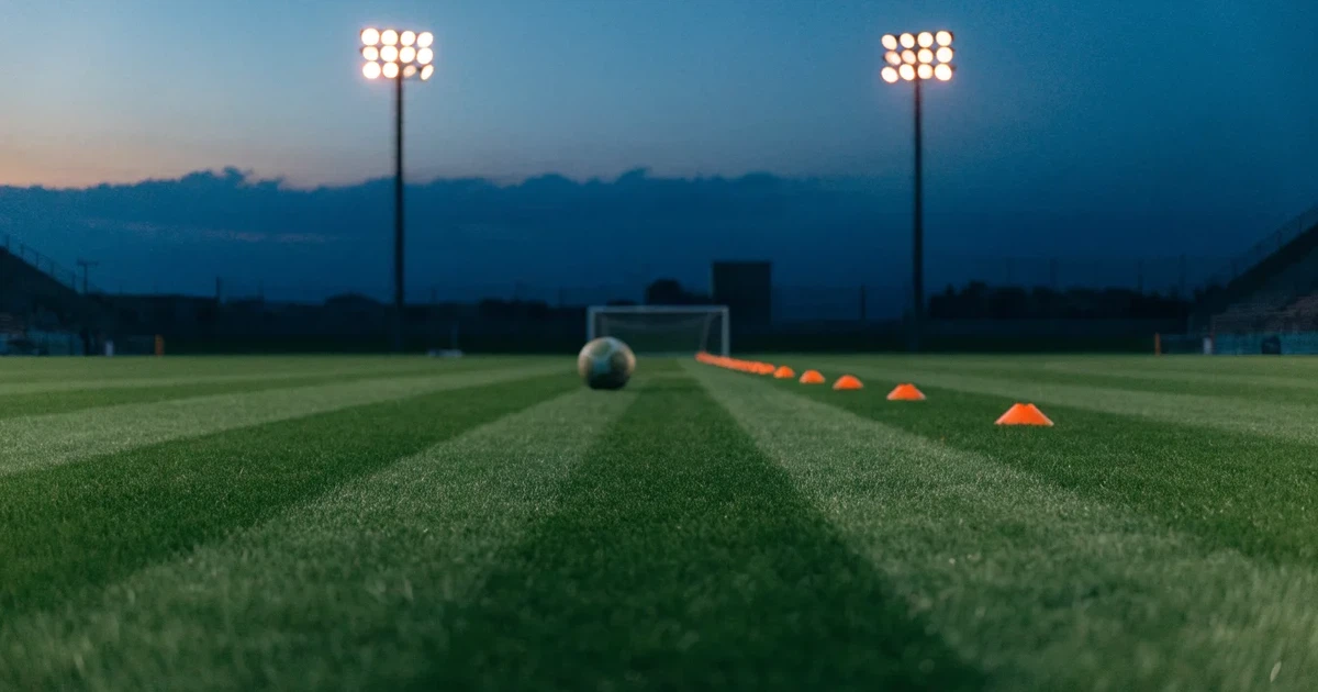 Eine Flutlichtanlage ragt über einen Trainingsplatz, auf dem Rasen liegen verstreut Hütchen und ein Ball, am Himmel die Abenddämmerung.