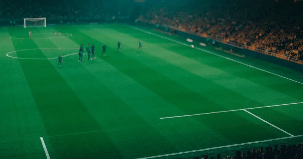 Ein großes Stadion bei Nacht, aus der Ferne unter Flutlicht gesehen, die Ränge hell erleuchtet, der Rasen leuchtend grün.