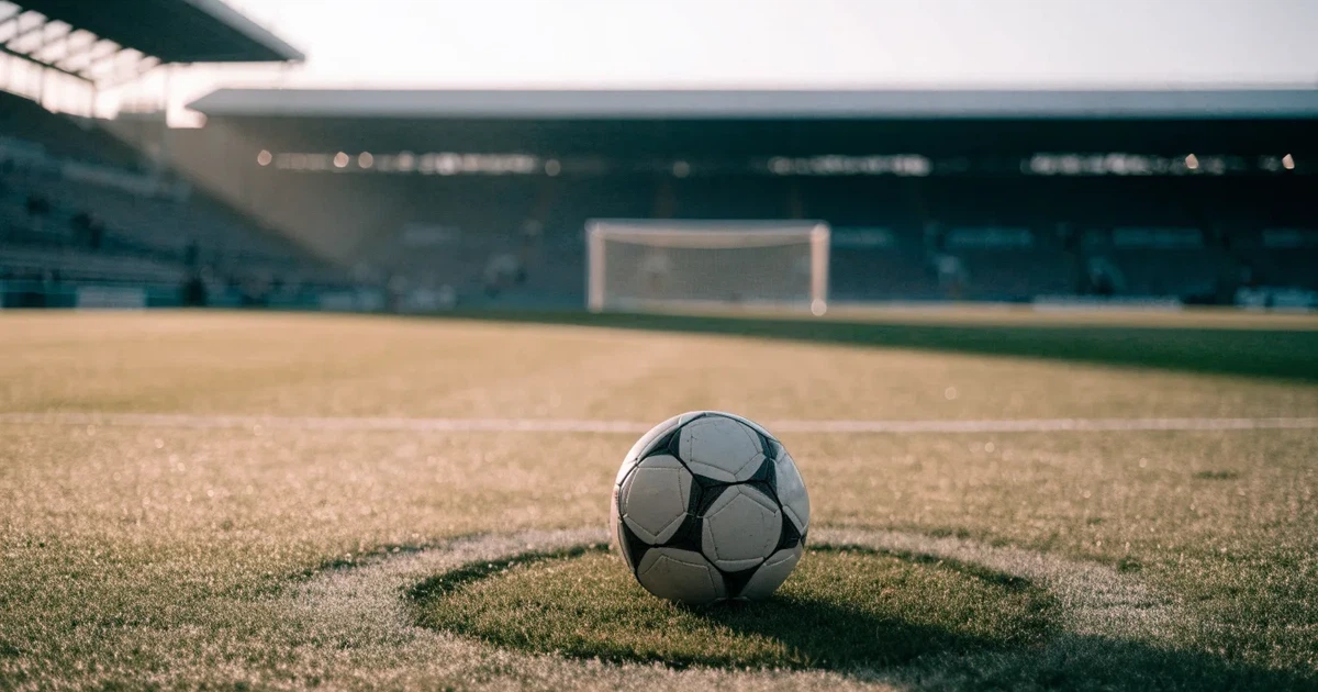 Ein Fußball liegt auf dem Mittelkreis eines Stadionrasens am frühen Morgen, im Hintergrund leere Ränge ohne erkennbare Beschriftung.