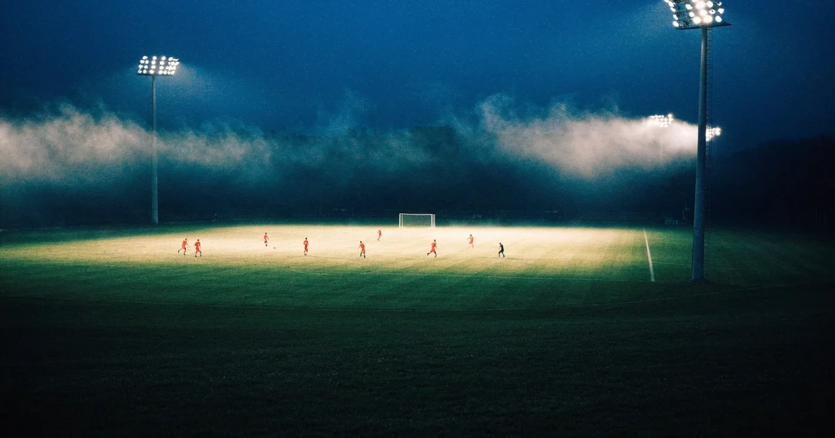 Ein Stadion unter Flutlicht, aus großer Entfernung über eine dunkle Wiese fotografiert, am Himmel zieht Nebel auf.