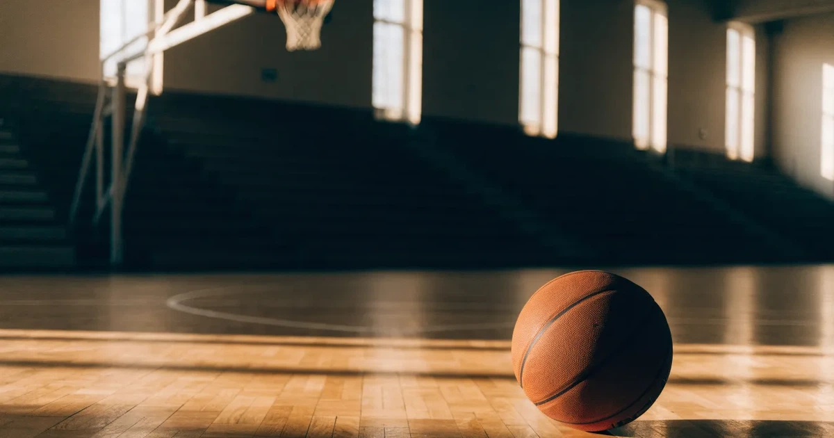 Ein Basketball liegt unter dem Korb auf dem Parkett einer leeren Sporthalle, Licht fällt durch hohe Fenster.