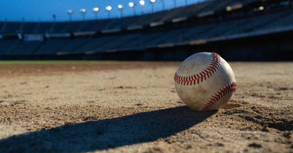 Ein Baseball liegt auf dem hellen Sand des Infields, im Hintergrund eine große Tribüne in der Abenddämmerung.