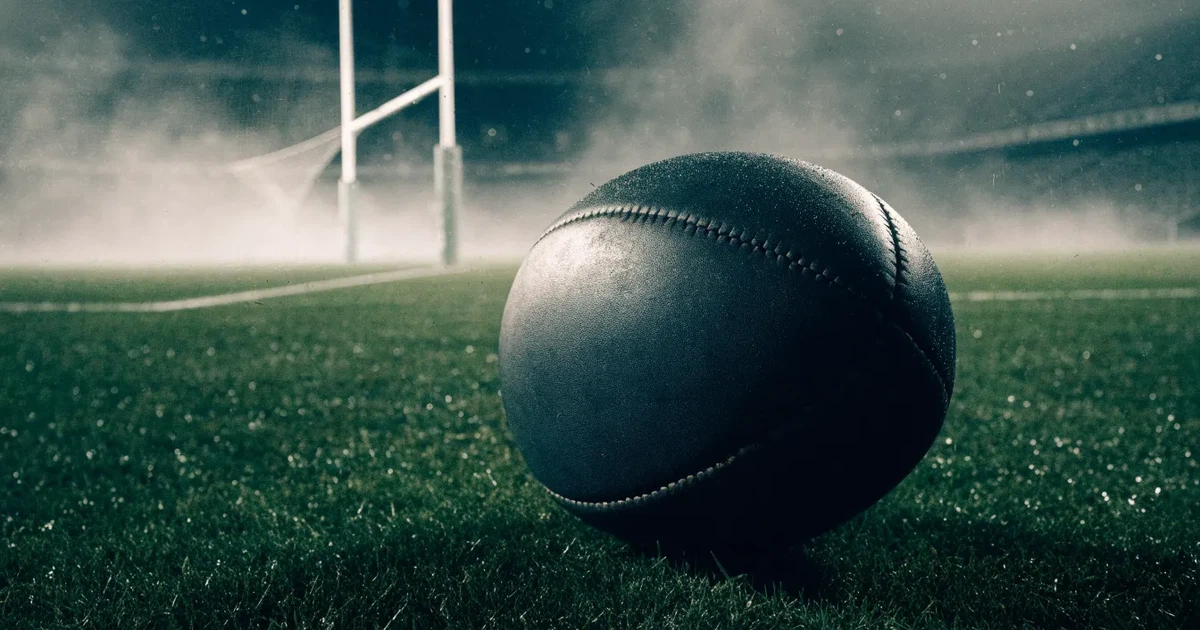 Ein ovaler Rugbyball liegt vor den hohen Torstangen auf nassem Rasen, über dem Stadion zieht Nebel auf.