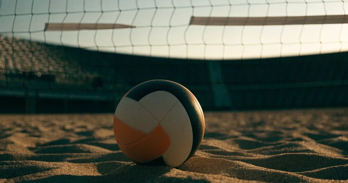 Ein Volleyball liegt auf dem Sand eines Beachvolleyball-Feldes neben dem gespannten Netz, im Hintergrund eine leere Tribüne bei Abendlicht.