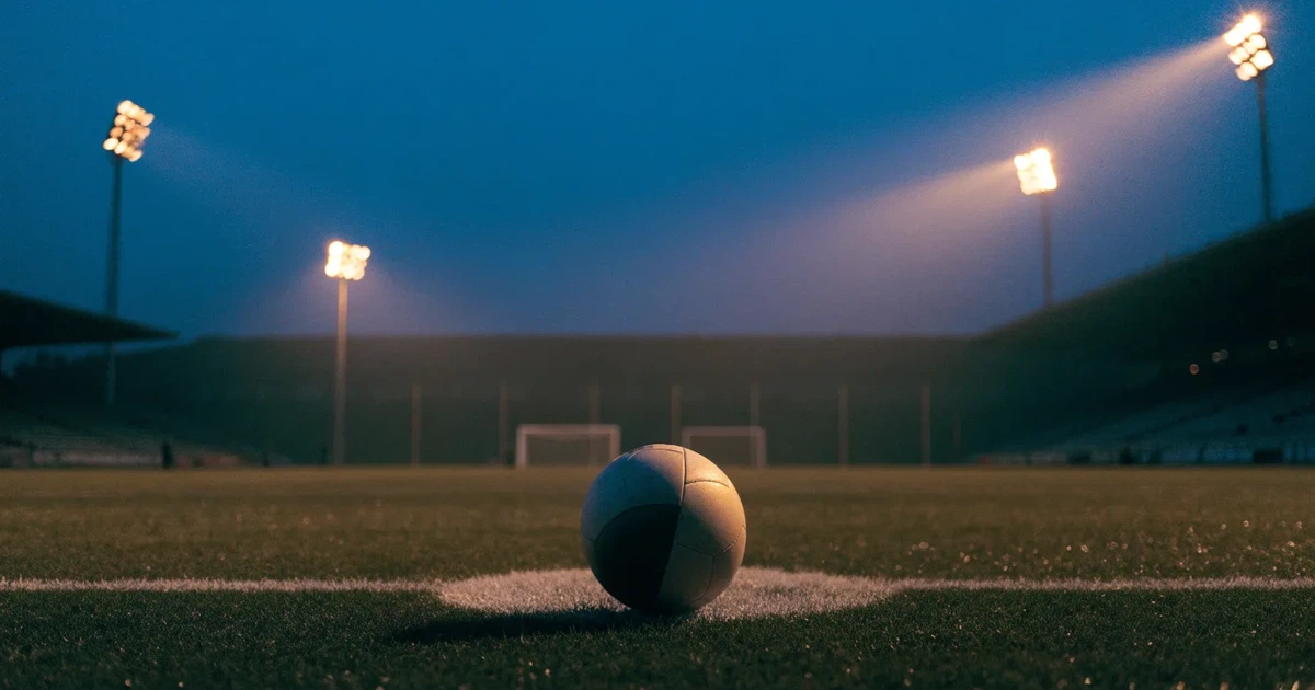 Ein Rasenplatz in der Dämmerung mit eingeschalteten Flutlichtmasten, ein Ball liegt am Anstoßpunkt, die Tribünen im Hintergrund sind unscharf.