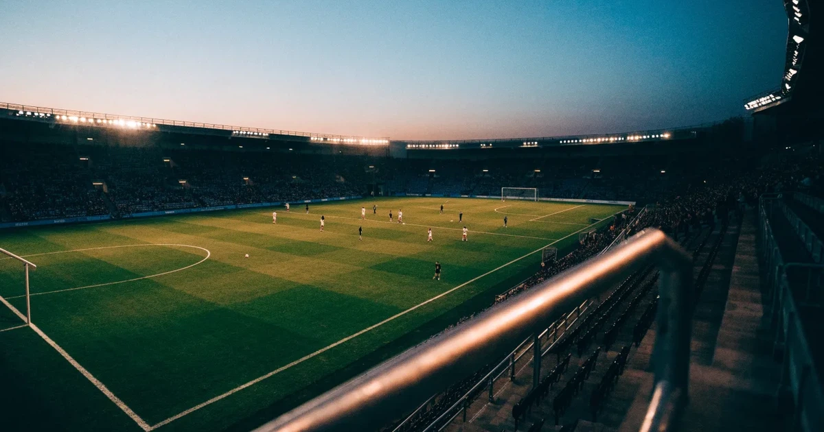 Ein Fußballstadion in der Dämmerung aus der Ferne, die Flutlichter gerade eingeschaltet, davor ein Kreisverkehr mit Laternen.